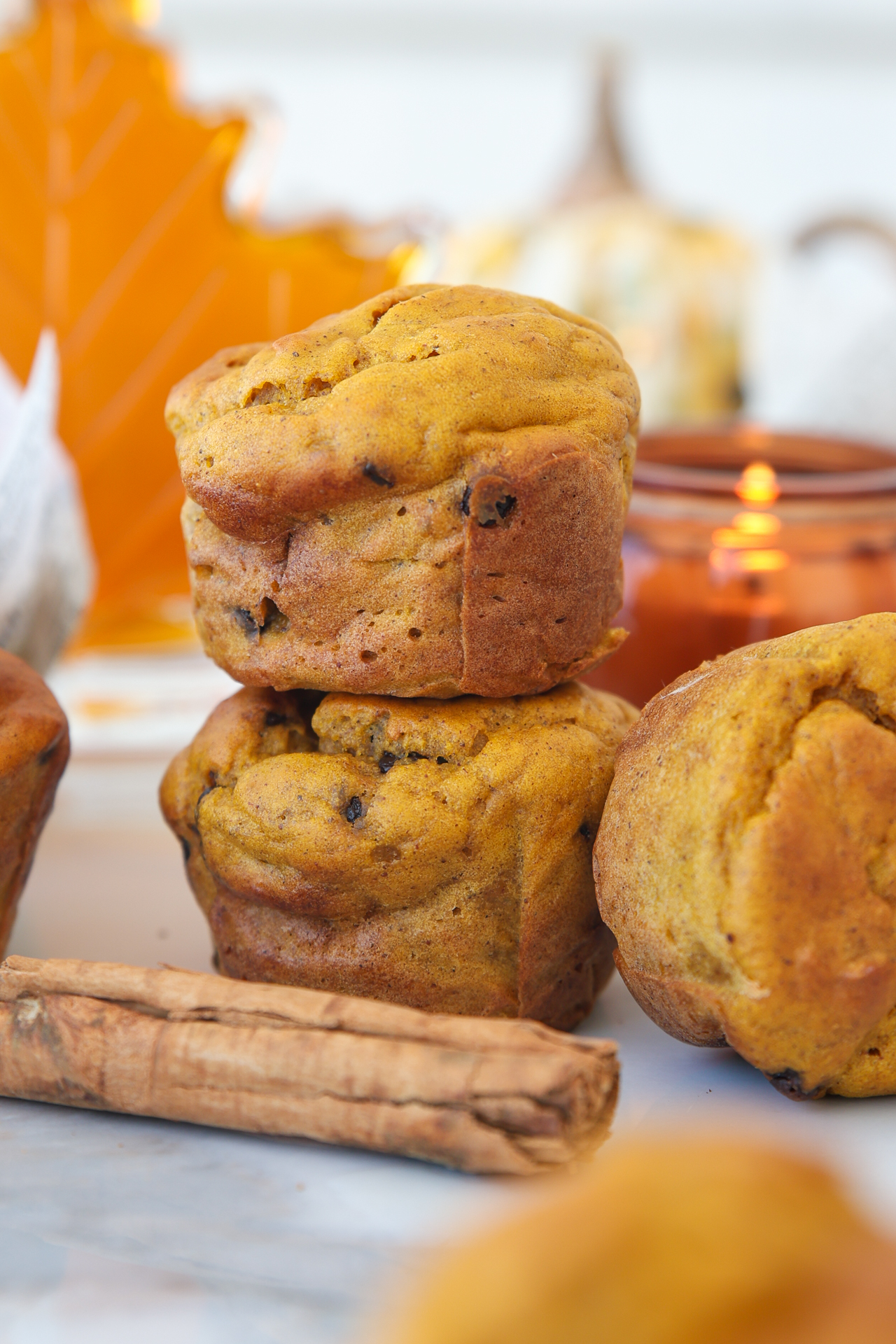 Two cottage cheese pumpkin muffins stacked, with a third muffin beside them. A cinnamon stick lies in front. In the background, there are orange autumn leaves, a candle, and a small pumpkin, creating a cozy fall atmosphere.