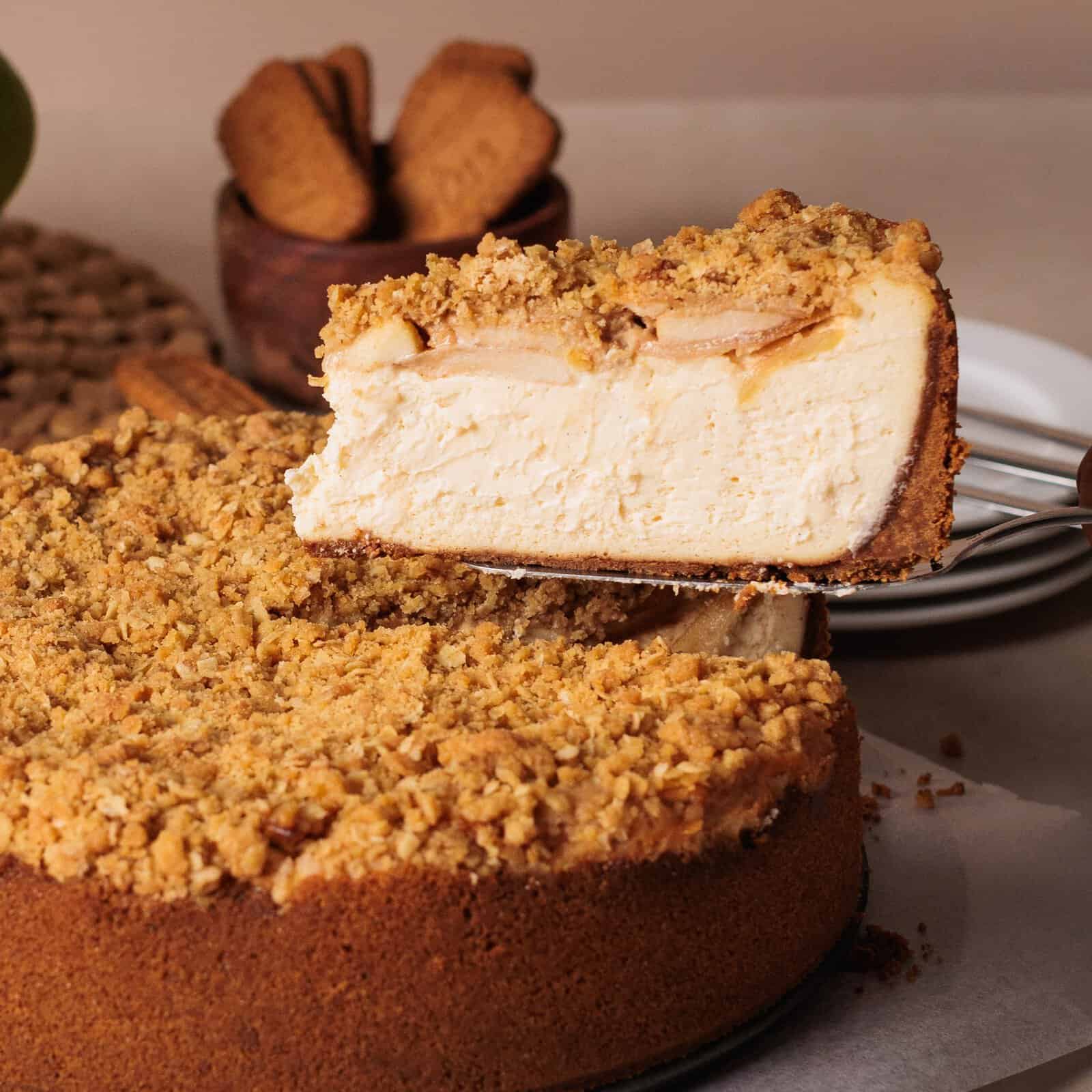 A metal server lifts a slice of apple crumble cheesecake away from the whole pie. The cheesecake has a thick, golden-brown crust, a creamy filling with apple slices, and a generous oat-based crumble topping. Blurred cookies sit in the background.