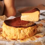 A slice of creamy, golden-brown cheesecake is lifted from a whole cheesecake resting on parchment paper, with plates in the background.