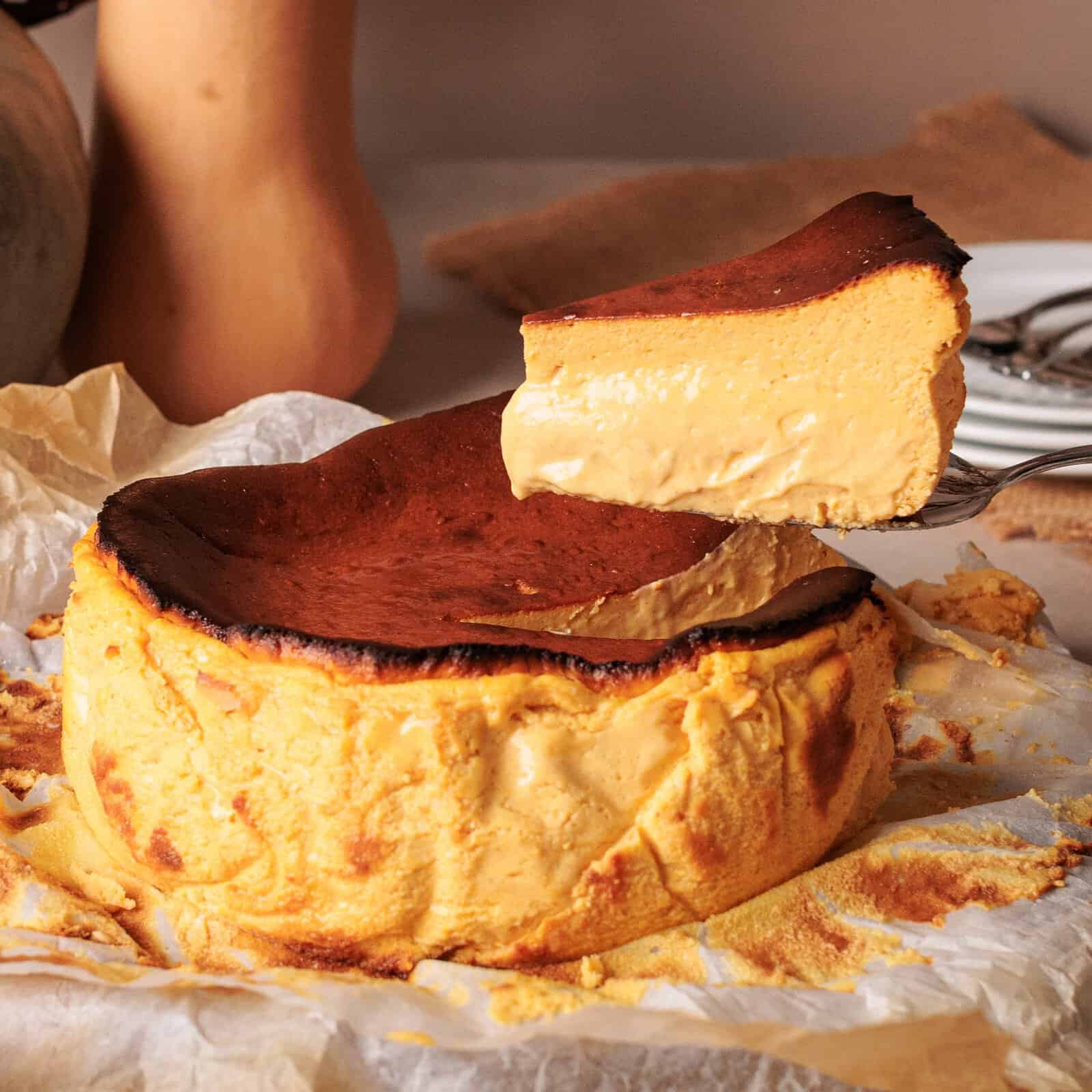 A slice of creamy, golden-brown cheesecake is lifted from a whole cheesecake resting on parchment paper, with plates in the background.