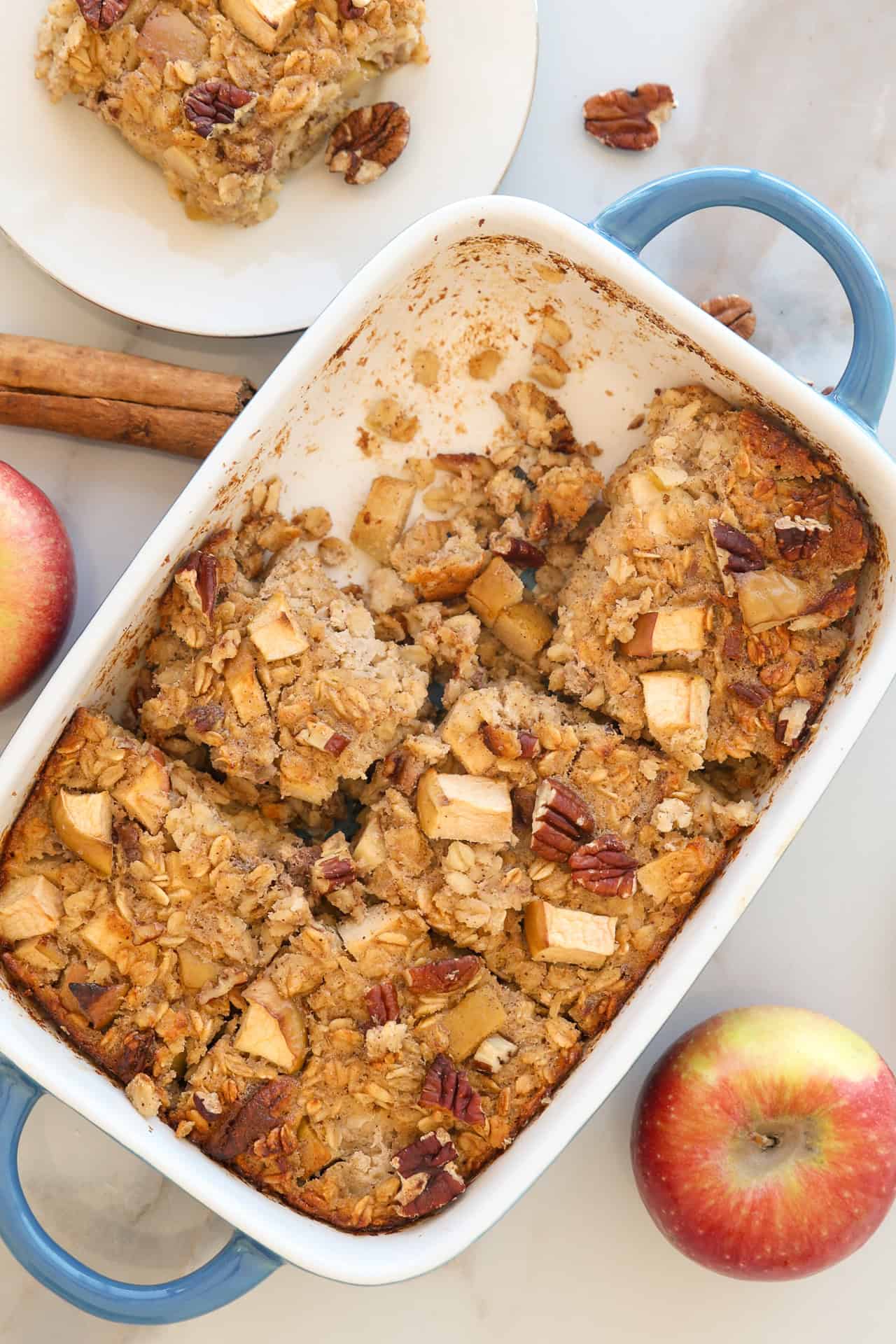 An apple cinnamon Baked Oatmeal with apple chunks and pecans, cut into squares in a white casserole dish with blue handles. Two apples, a cinnamon stick, and a plate with a serving are nearby.