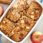 Apple cinnamon Baked Oatmeal with diced apples and pecans in a white dish, partially sliced. Fresh apples and cinnamon sticks are nearby on the table.
