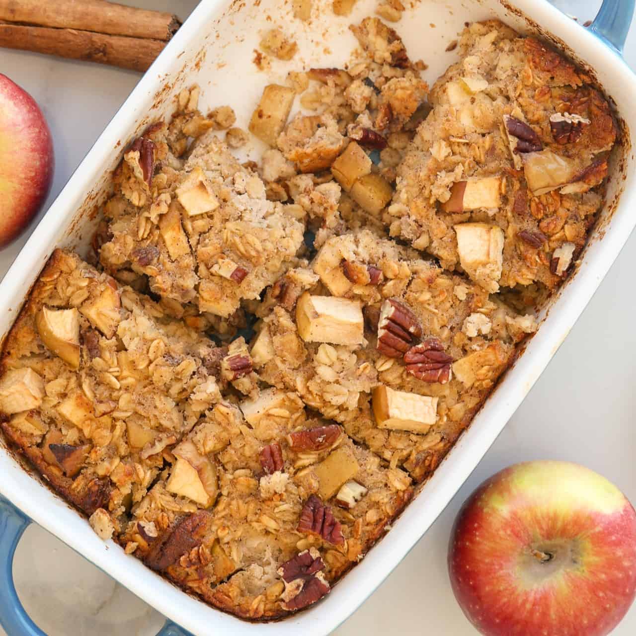 Apple cinnamon Baked Oatmeal with diced apples and pecans in a white dish, partially sliced. Fresh apples and cinnamon sticks are nearby on the table.