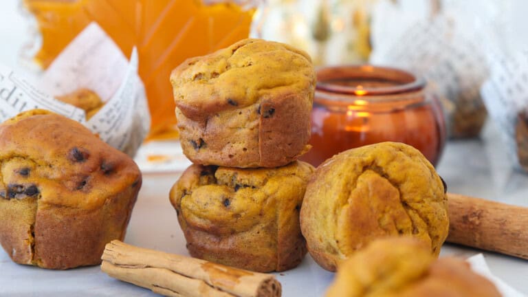 Cottage cheese pumpkin muffins with chocolate chips are stacked on a table, surrounded by cinnamon sticks, a brown candle holder, and a glass jar of orange syrup or honey in the background.