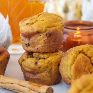 Three cottage cheese pumpkin muffins are stacked beside a cinnamon stick, with an orange candle and pumpkins blurred in the background. The muffins have a golden-brown color and a rustic, homemade appearance.