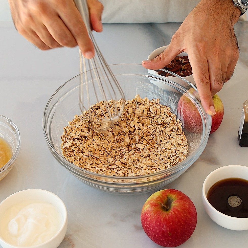 A person whisks oats in a glass bowl on a table with apples, yogurt, syrup, and a small bowl of pecans nearby, preparing ingredients for a recipe.