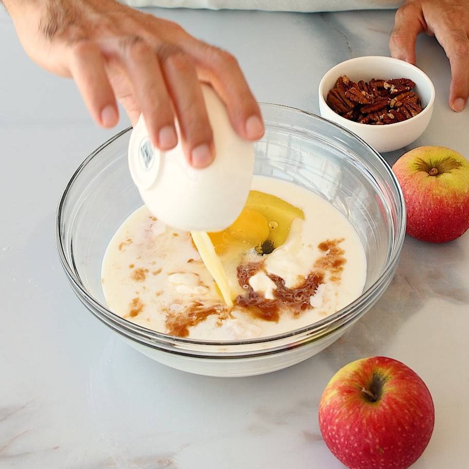 A hand pours liquid from a small white cup into a bowl with eggs, milk, and vanilla. Apples and a bowl of pecans are on the counter nearby.