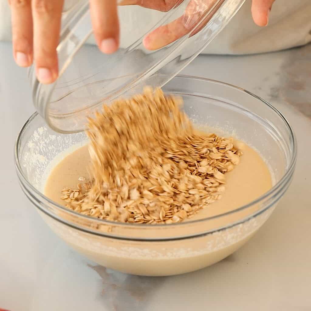 A hand pours rolled oats from a glass bowl into another bowl filled with a creamy batter, preparing ingredients for baking on a light countertop.