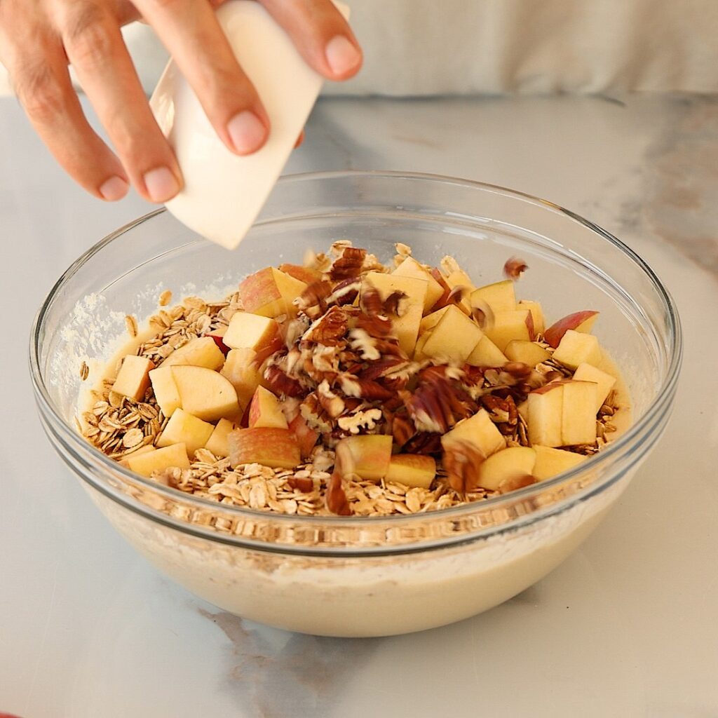 A hand pours chopped pecans into a glass bowl filled with oats, apple chunks, and batter, preparing a mixture on a light countertop.