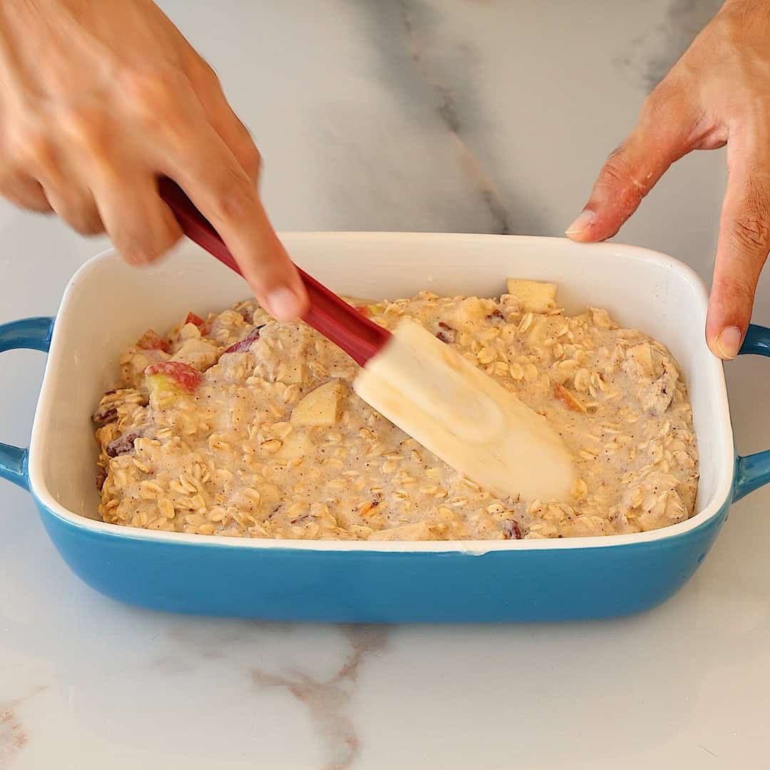 Hands use a spatula to spread an oat and apple mixture evenly in a blue rectangular baking dish on a marble surface.