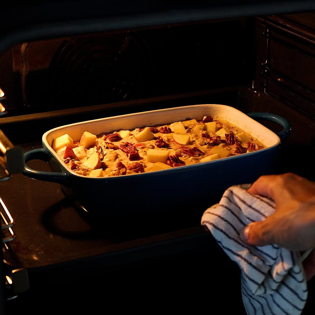 A hand holding a striped towel pulls a blue baking dish with baked oatmeal with apples and pecans out of an oven.