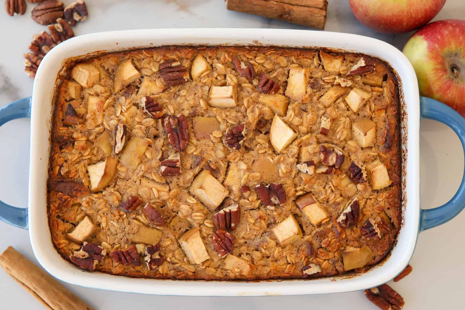 A baked oatmeal casserole topped with apple chunks and pecans sits in a blue-handled dish, surrounded by apples, cinnamon sticks, and pecans on a white surface.