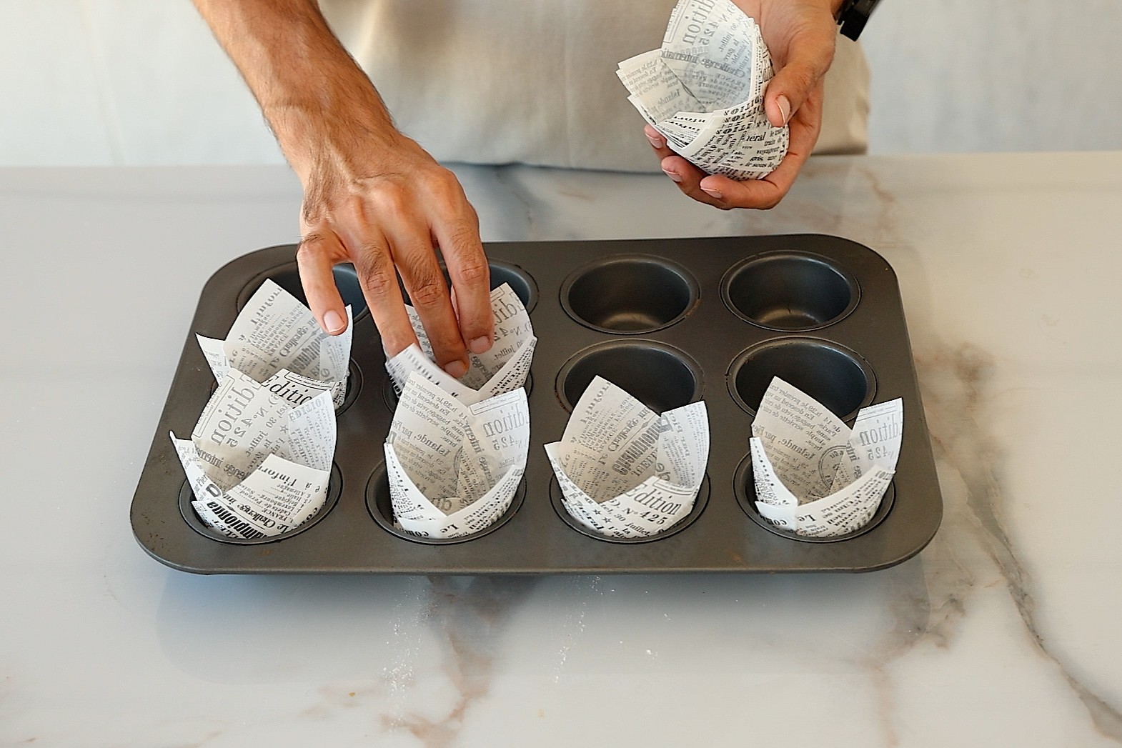 A person places decorative paper liners with printed text into a muffin tin on a marble countertop.