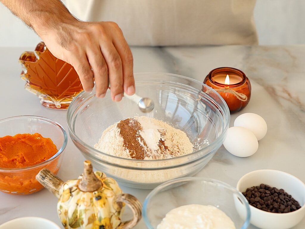 A person adds spices to a bowl of flour surrounded by eggs, pumpkin puree, chocolate chips, and other baking ingredients on a kitchen counter.