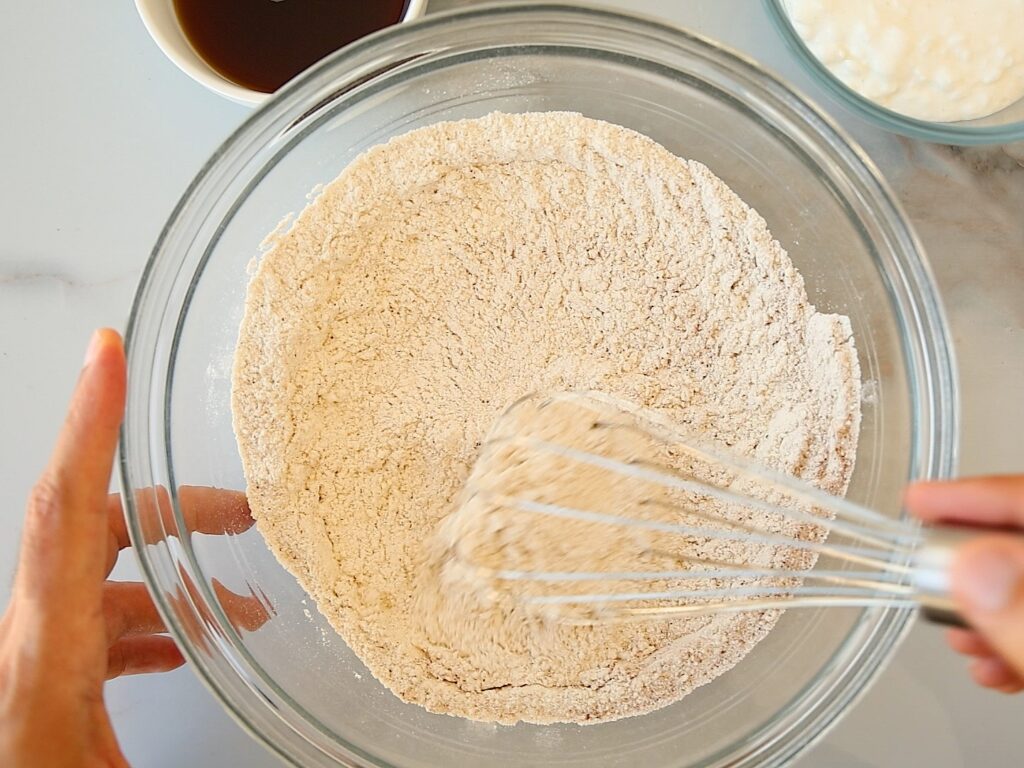 A person whisks flour in a clear glass bowl on a white surface, with two bowls of liquid ingredients nearby.