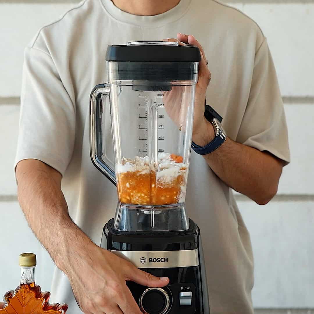 A person in a beige shirt uses a Bosch blender with cottage cheese, pumpkin puree, buttermilk and maple inside. Their left hand holds the lid, and their right turns the dial. A maple syrup bottle sits on the counter.