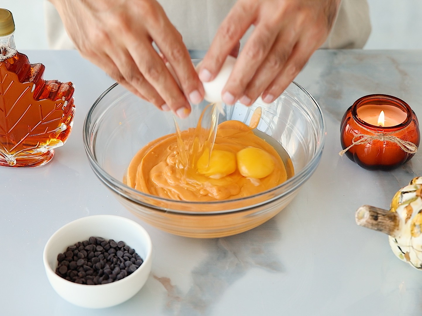 A person cracks eggs into a glass bowl of cottage cheese and pumpkin muffin batter on a marble counter, surrounded by chocolate chips, maple syrup, a lit candle, and a small decorative pumpkin.