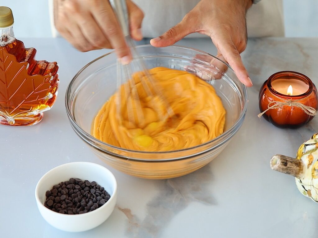 A person whisks a yellow batter in a clear bowl. Nearby are a leaf-shaped syrup bottle, a bowl of chocolate chips, a lit candle, and a small decorative pumpkin on a marble surface.