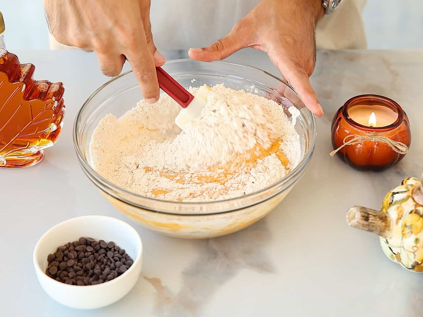 A person mixes flour into a bowl of batter with a red spatula. Nearby are a jar of maple syrup, a bowl of chocolate chips, a candle, and a decorative gourd on a light-colored countertop.