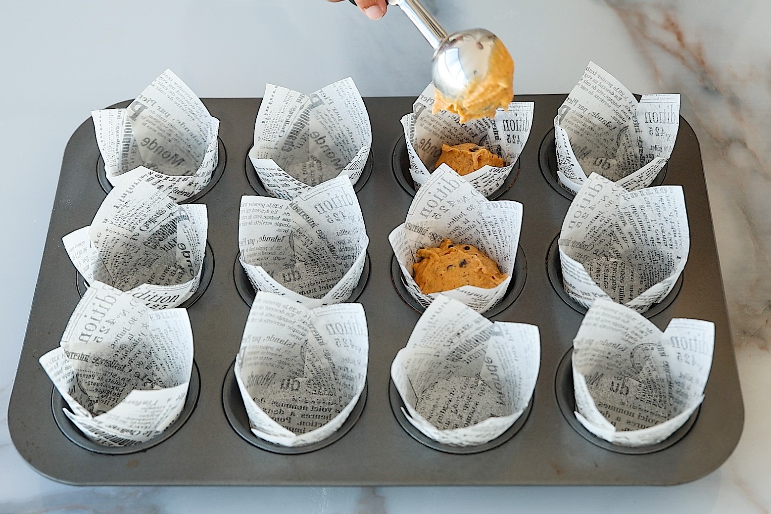 A hand scoops muffin batter into tulip-style muffin liners with printed text, placed in a 12-cup muffin tray on a marble surface.
