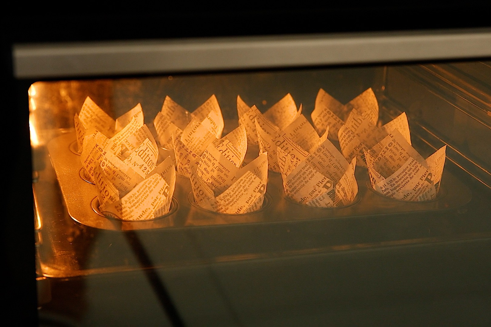 Cupcake liners with newspaper print are arranged in a muffin tin inside an oven, with the oven light on and the liners softly illuminated as they bake.