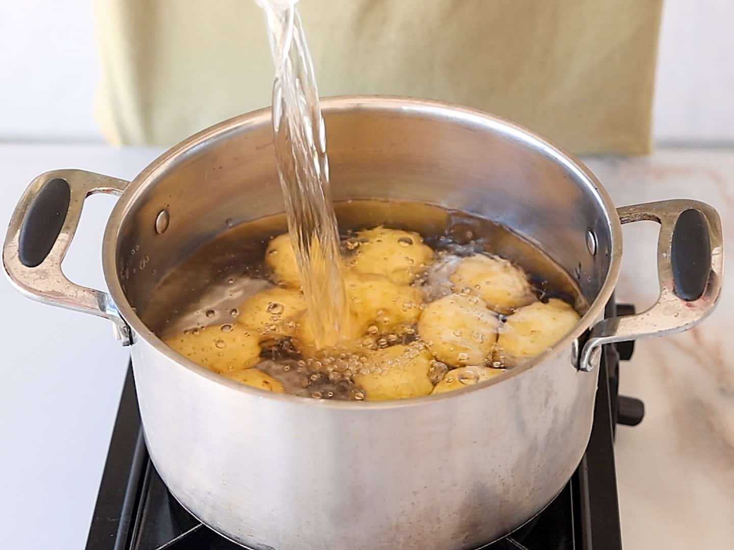 Water is being poured into a pot of boiling small baby potatoes on a stove, with someone in a yellow shirt standing behind.