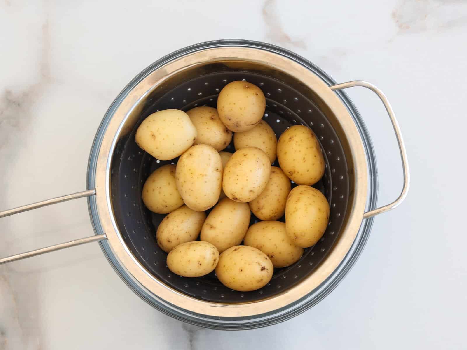 A metal colander holds several small, whole yellow potatoes that were boiled and are now drying, placed on a light marble surface.
