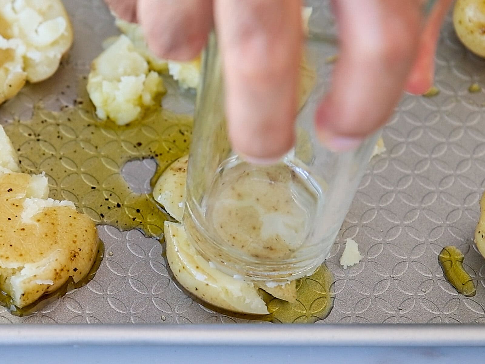 A hand uses a glass to smash boiled potatoes on a baking sheet coated with oil, preparing them for roasting.