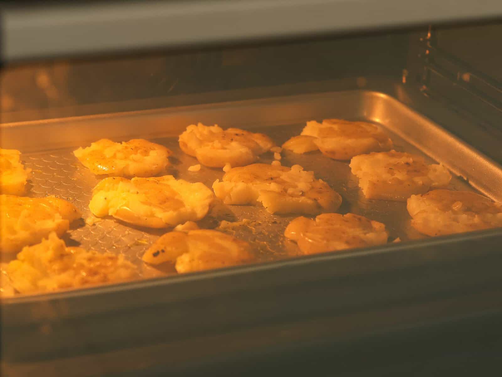A baking tray with several smashed potatoes is shown inside an oven, cooking. The potatoes appear golden and are spread out evenly on the tray under the warm oven light.