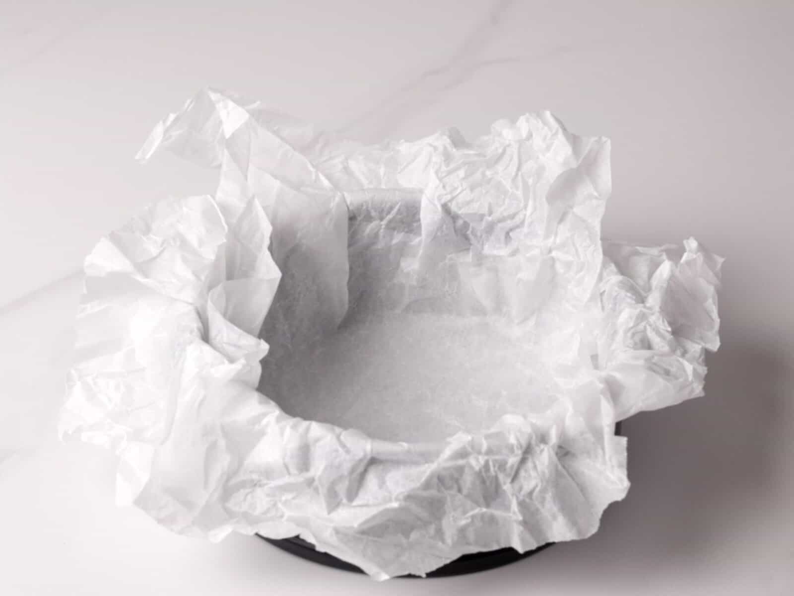 A round baking pan lined with crumpled parchment paper sits on a white surface, ready for use.