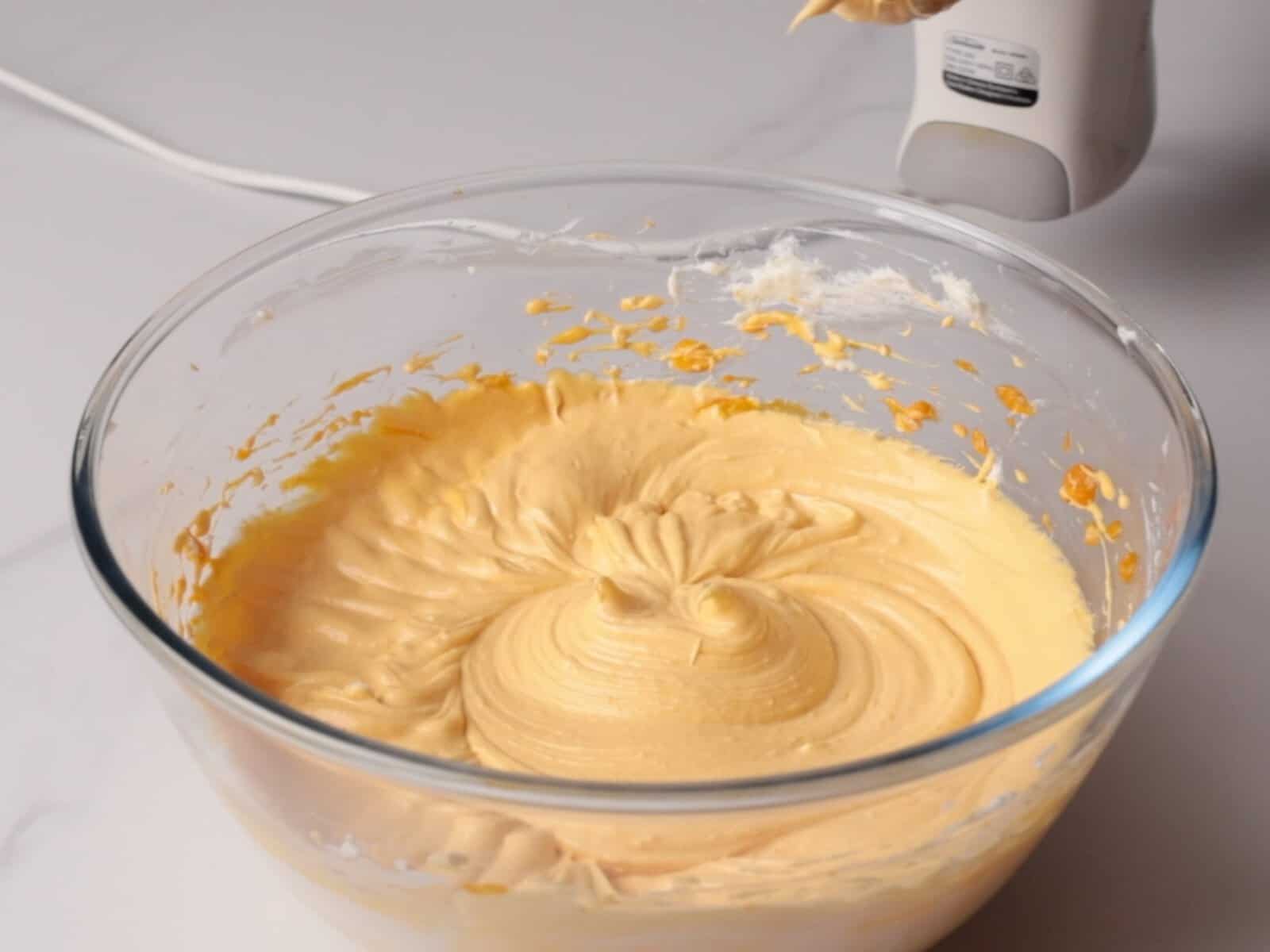 A glass bowl filled with creamy, pumpkin basque cheesecake batter sits on a white surface beside a hand mixer with batter on its beater.