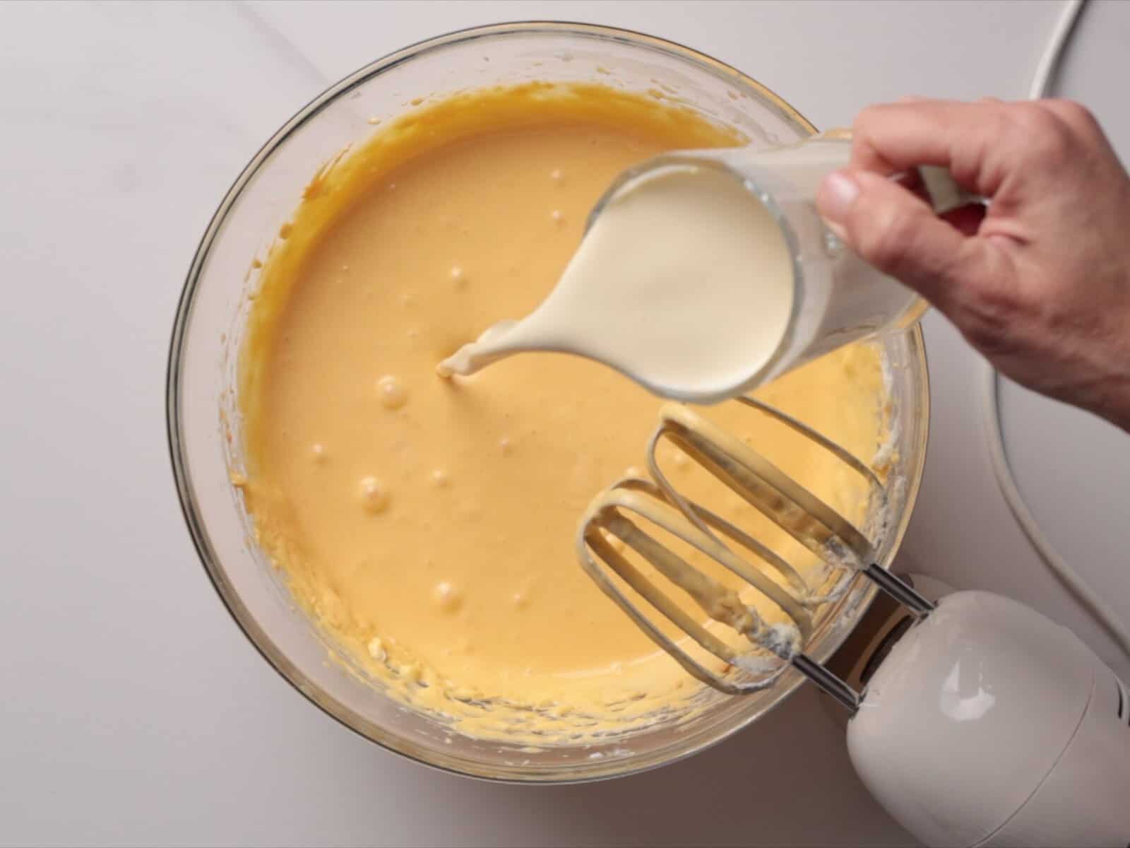 A hand pours cream from a glass pitcher into a bowl of pumpkin cheesecake batter next to an electric hand mixer on a white surface.