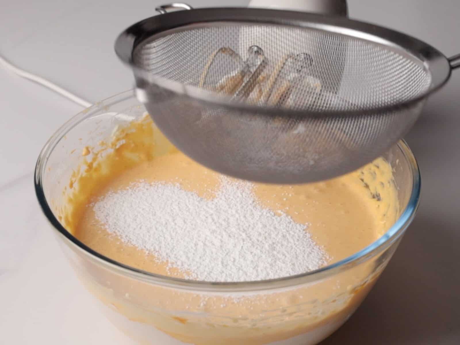 A metal sifter is positioned over a bowl of pumpkin cheesecake batter, with flour being sifted into the mixture. An electric mixer is visible in the background.