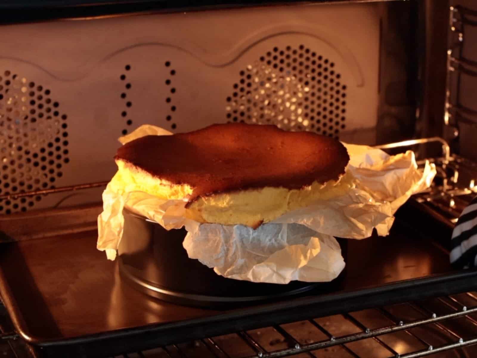 A pumpkin basque heesecake with a golden brown top sits in a parchment-lined pan on an oven rack, baking.