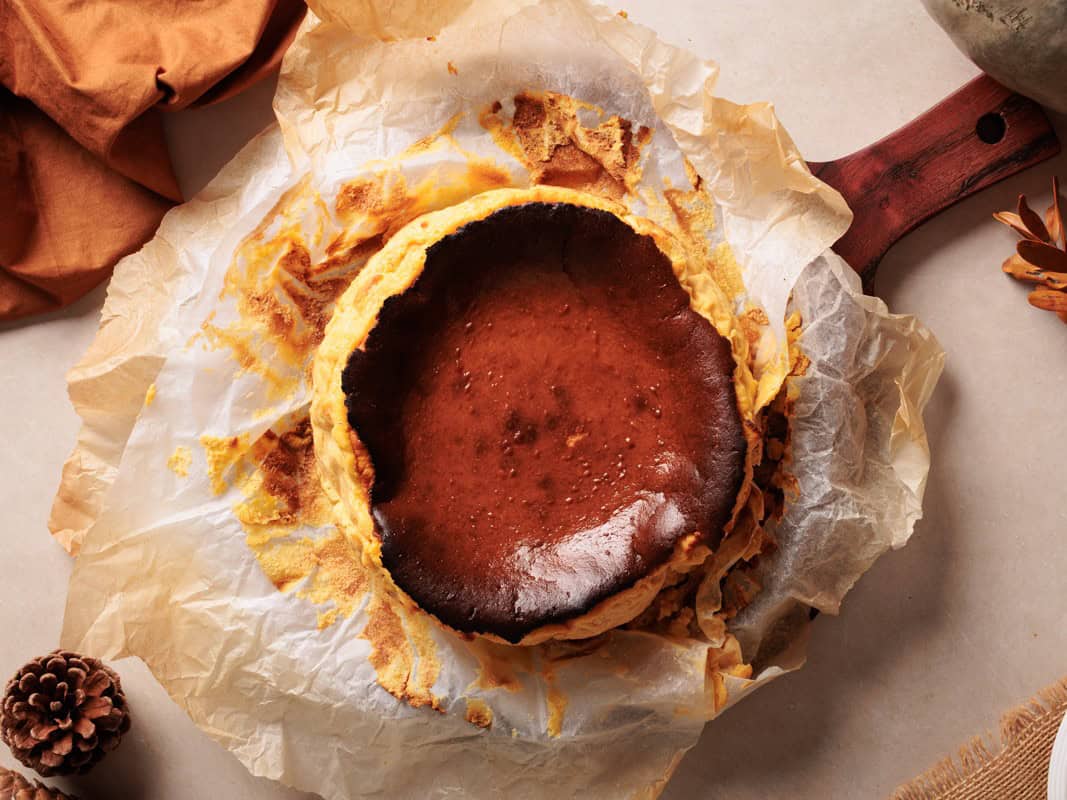 A crustless burnt Basque cheesecake sits on crumpled parchment paper atop a wooden board, surrounded by autumn decor like pinecones and a brown cloth.