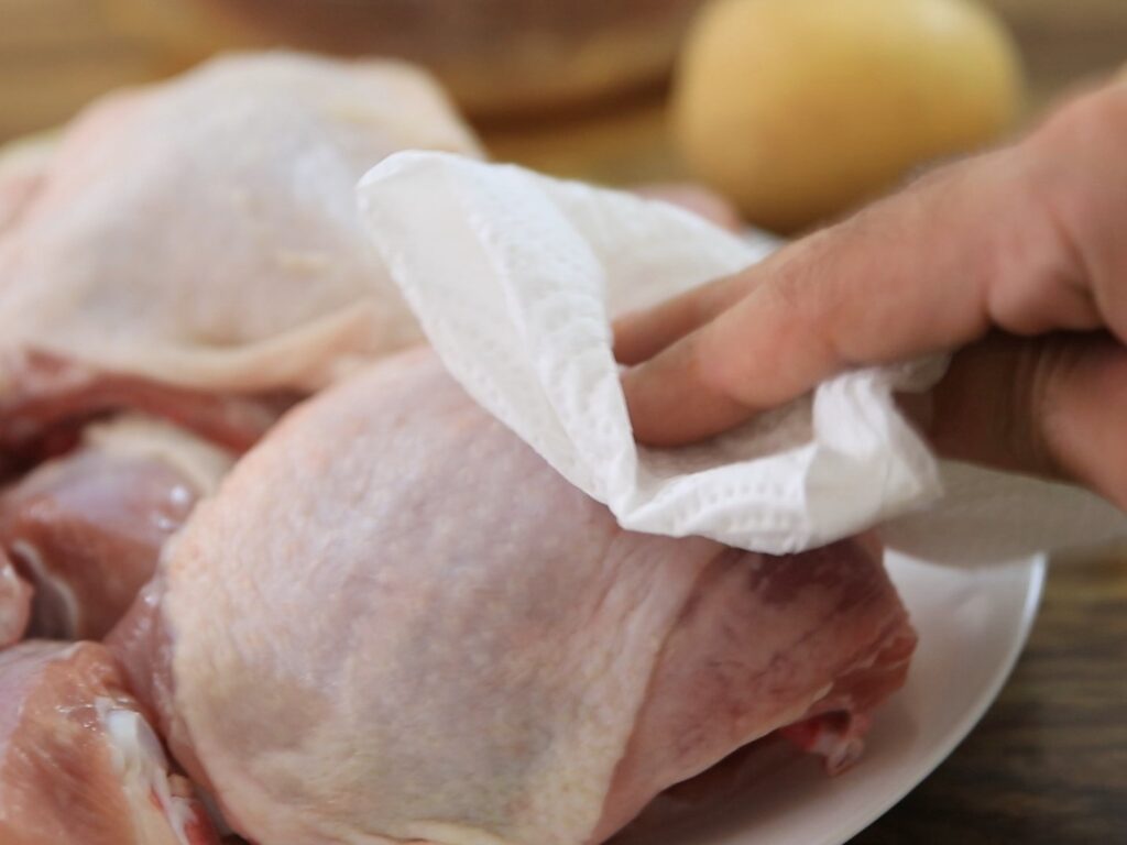 A hand uses a paper towel to pat dry raw chicken pieces on a white plate, with a blurred potato in the background.
