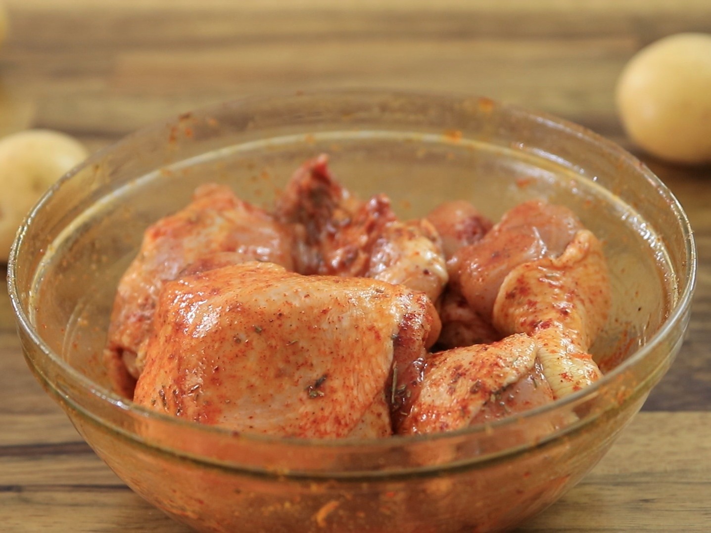 Raw chicken pieces coated with a reddish spice marinade in a clear glass bowl, placed on a wooden surface with blurred potatoes in the background.