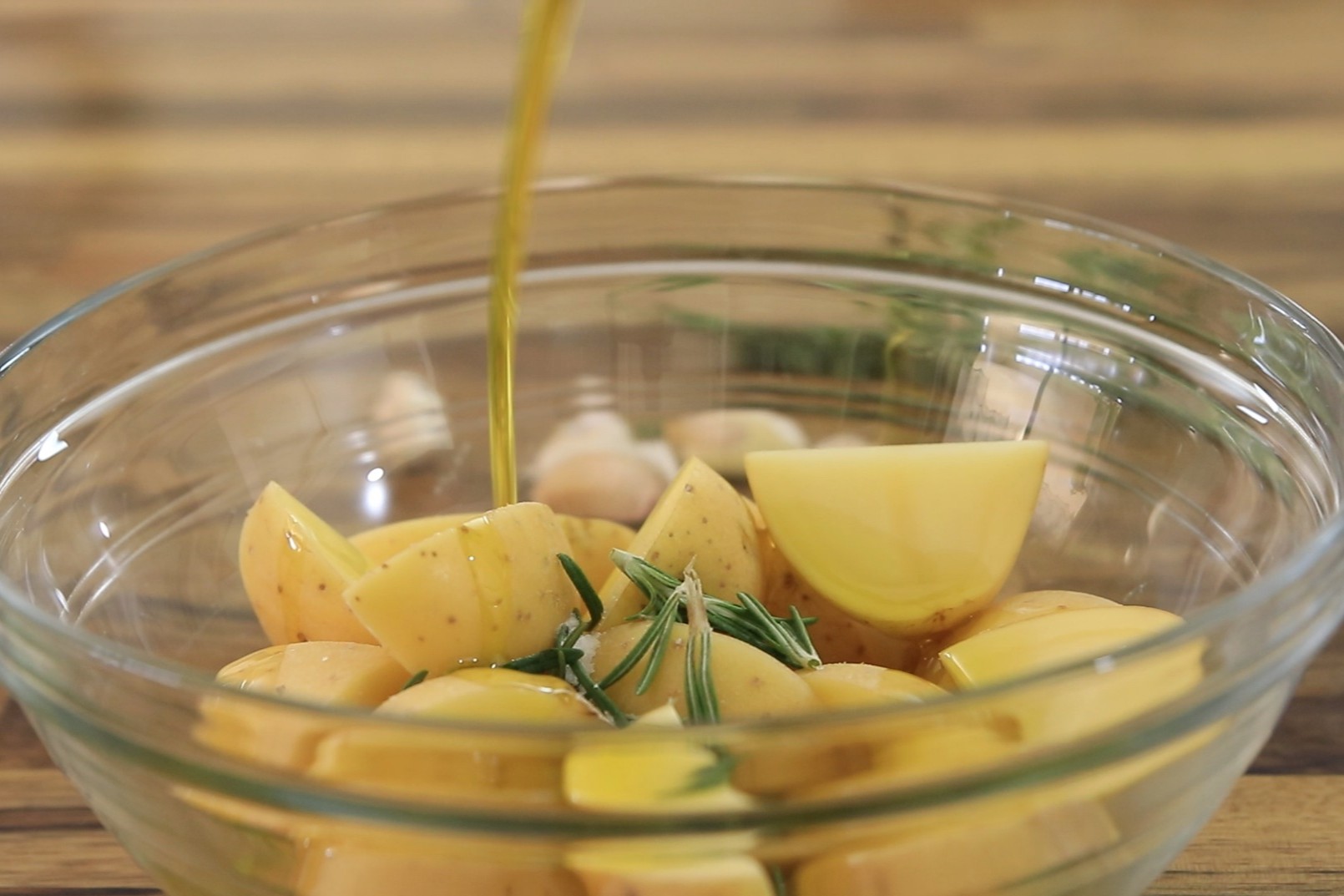 Olive oil is being poured over quartered yellow potatoes and rosemary sprigs in a clear glass bowl, with a wooden surface in the background.
