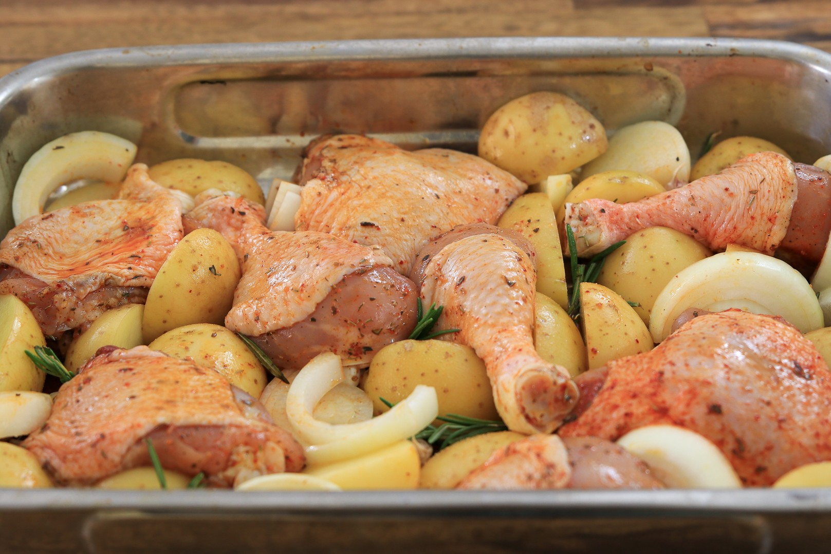 Raw chicken pieces, halved potatoes, onion slices, and herbs are arranged in a metal roasting pan, ready to be cooked.