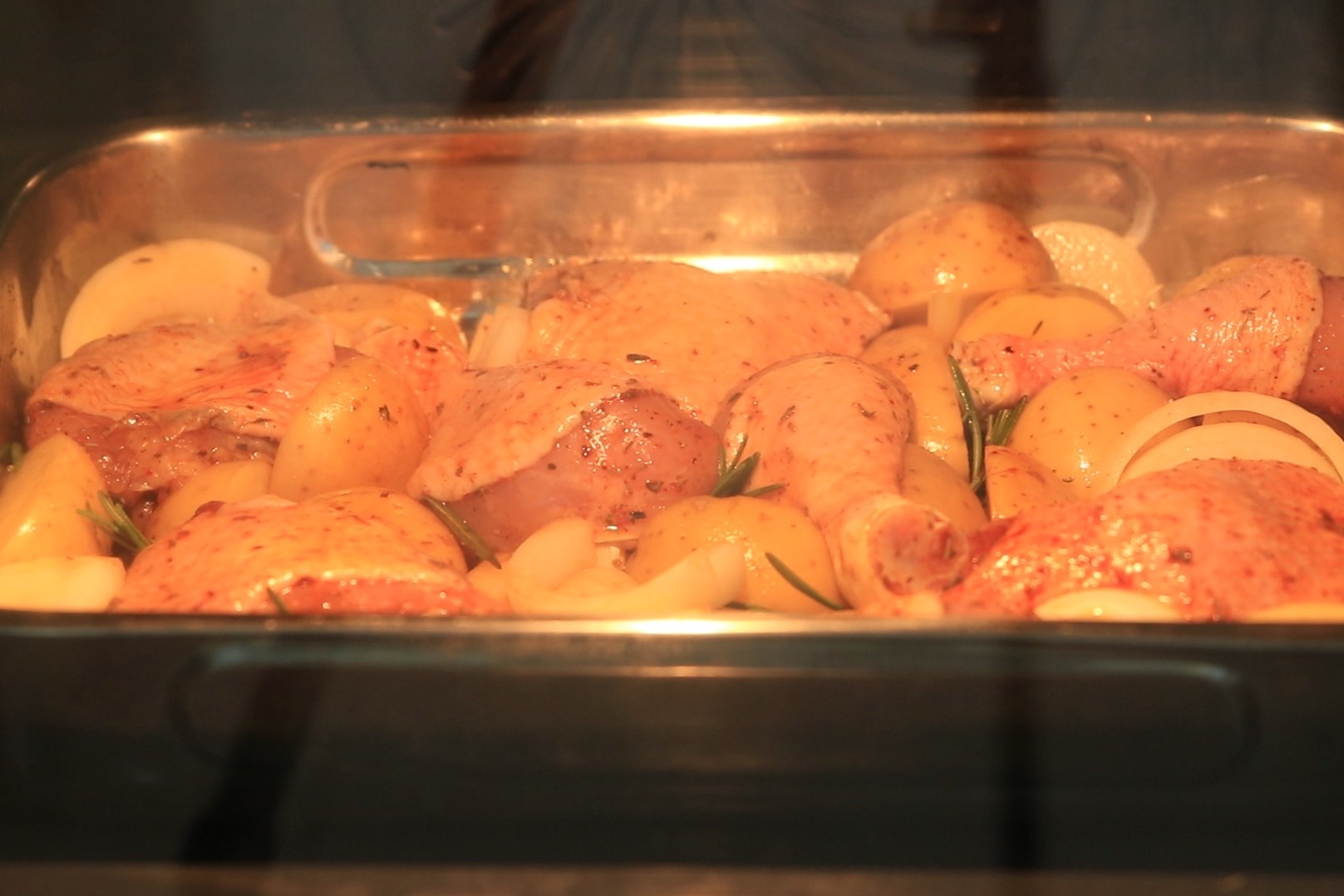 Chicken pieces, potatoes, and onions roasting in a metal baking tray inside an oven. The food is seasoned and partially browned, with steam visible on the oven door.