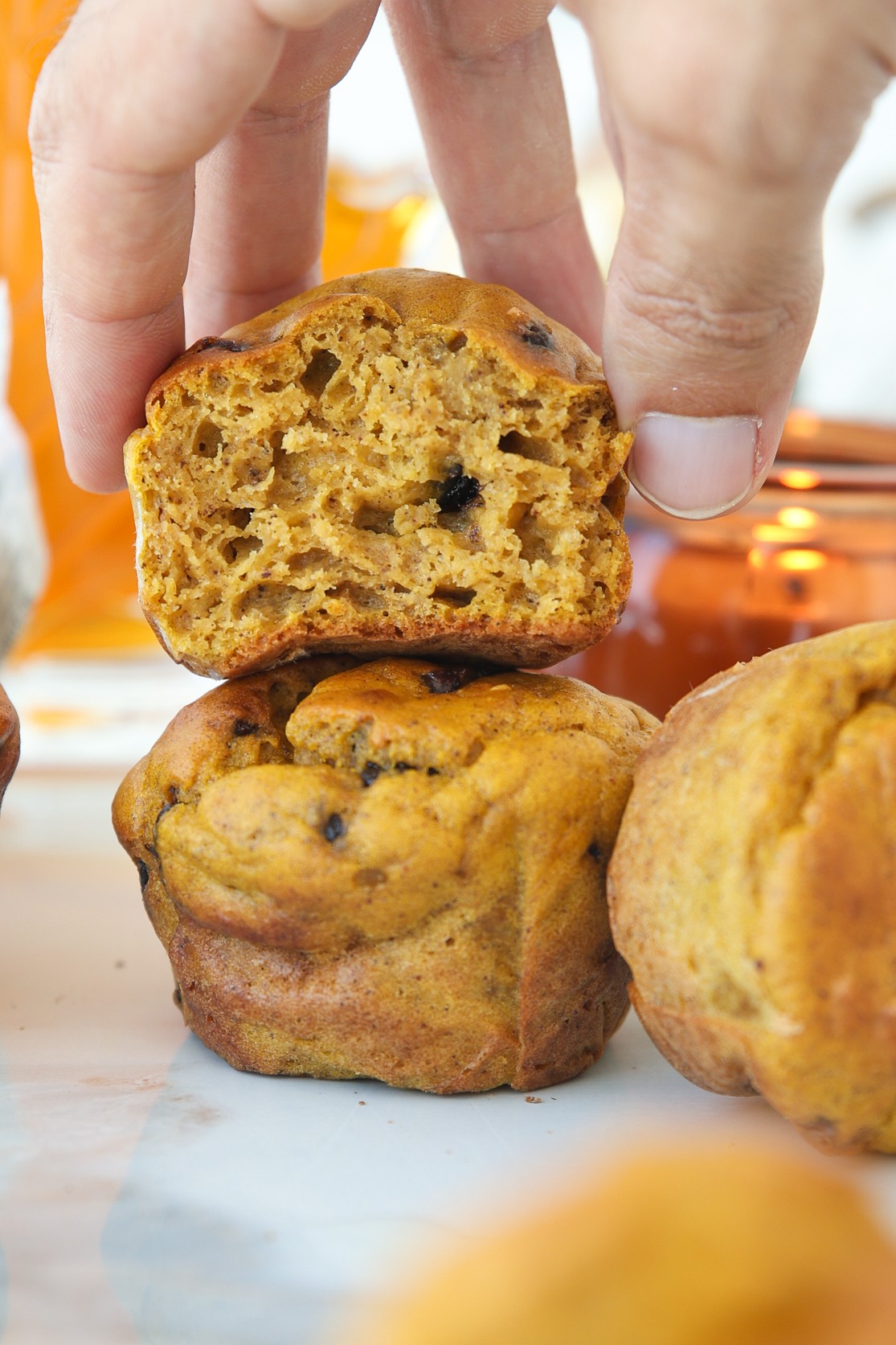 A hand holds a halved pumpkin cottage cheese muffin with chocolate chips above two whole muffins, showing its moist, fluffy texture.