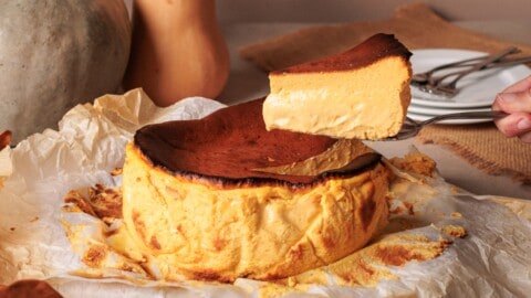 A hand lifts a slice of creamy, golden pumpkin Basque burnt cheesecake from the whole cake, which sits on parchment paper with a caramelized top and rich, soft interior.
