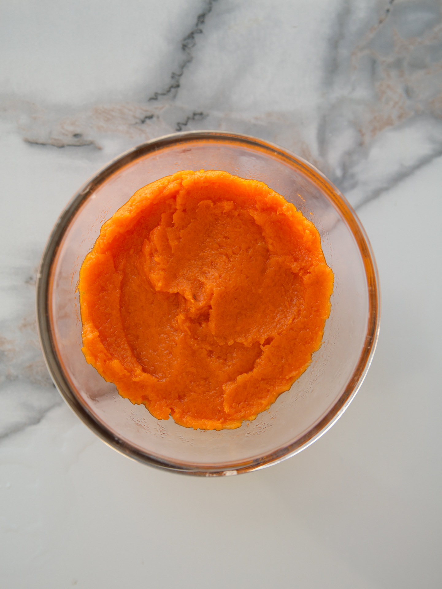 A clear glass bowl filled with smooth, bright pumpkin puree sits on a white and gray marble surface.