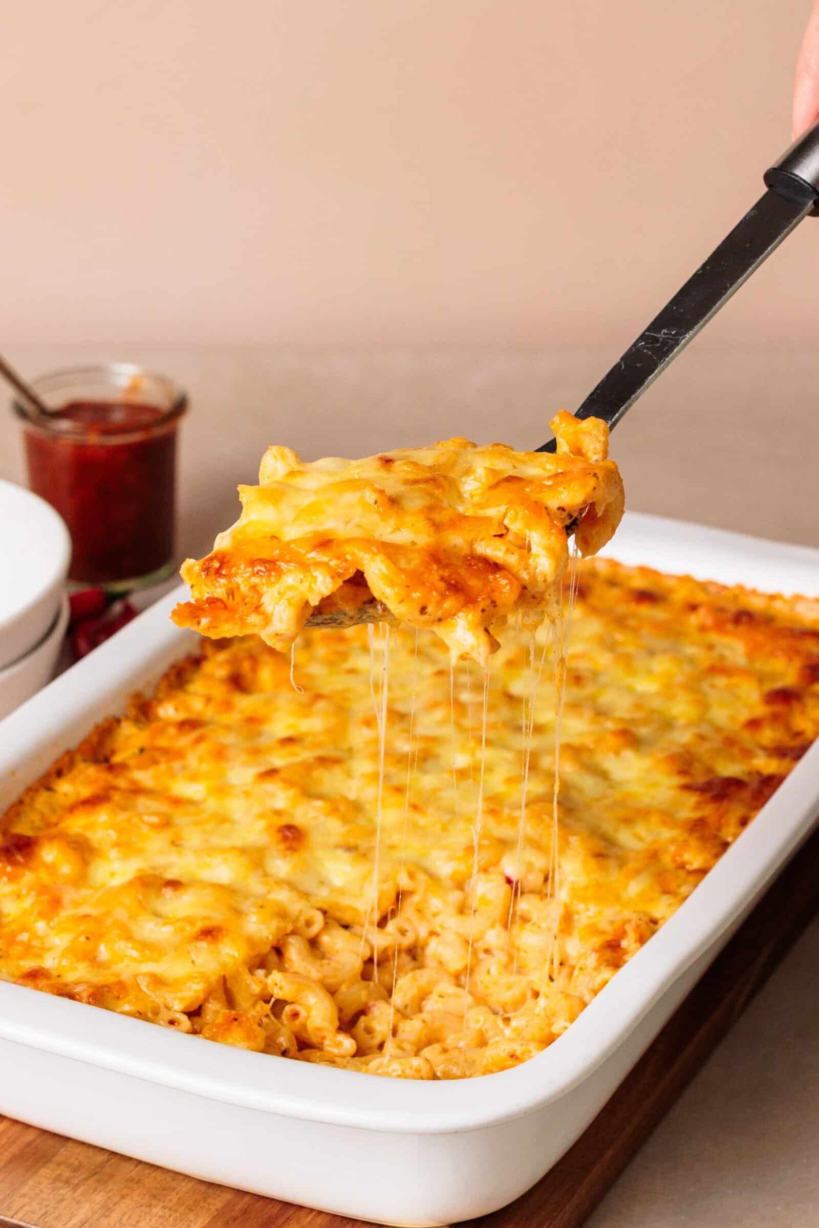 A spatula lifts a cheesy portion of baked macaroni and cheese from a white casserole dish, with gooey melted cheese stretching from the pan.
