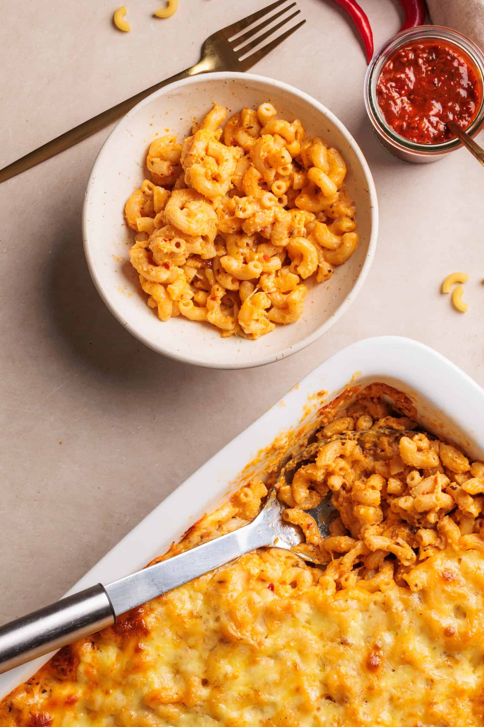 A bowl of baked spicy macaroni and cheese sits beside a casserole dish filled with more macaroni. A serving spoon rests in the dish. A jar of harissa paste and utensils are nearby on a beige surface.