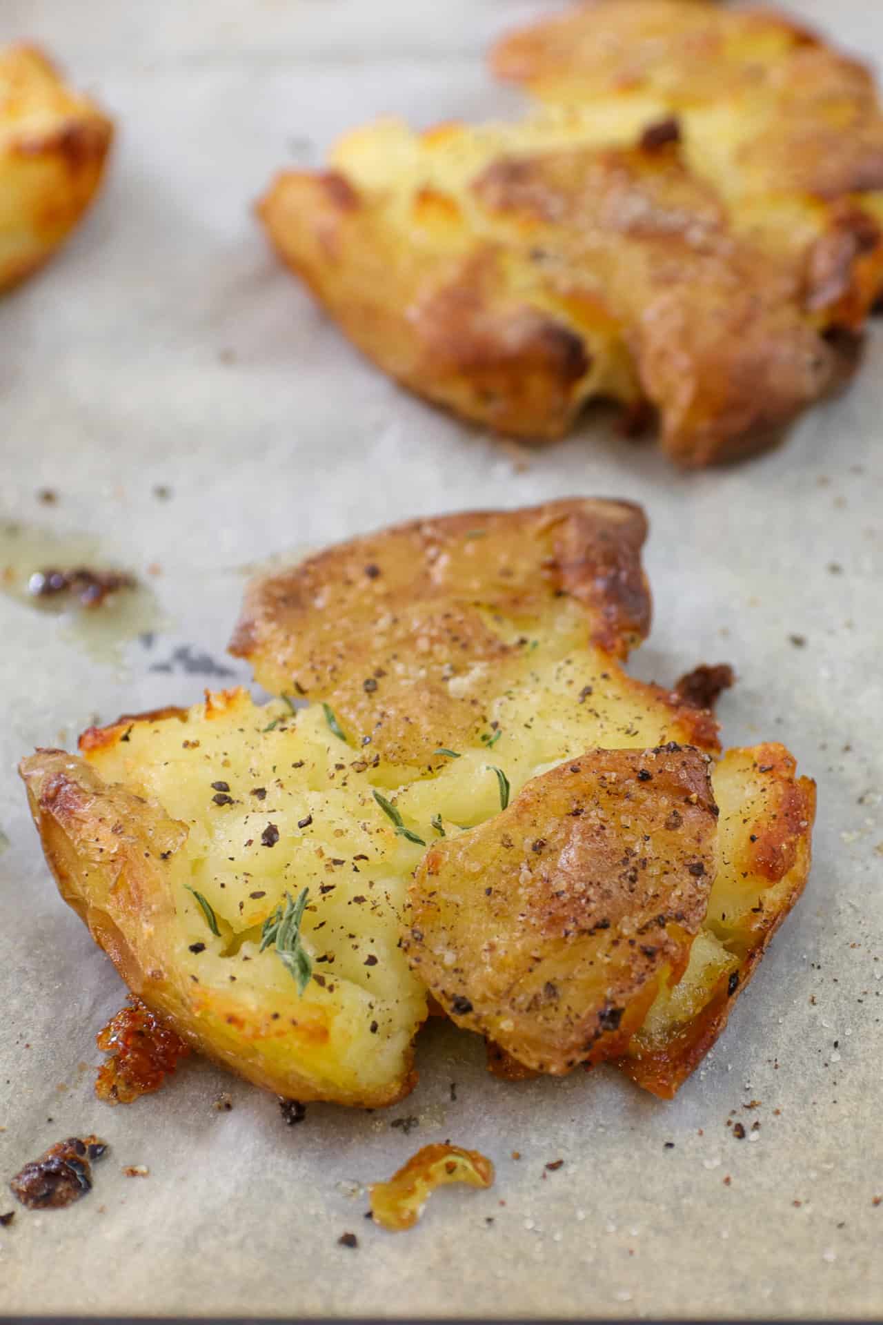 Crispy smashed potatoes topped with cracked black pepper and herbs sit on parchment paper, with another potato in the background.