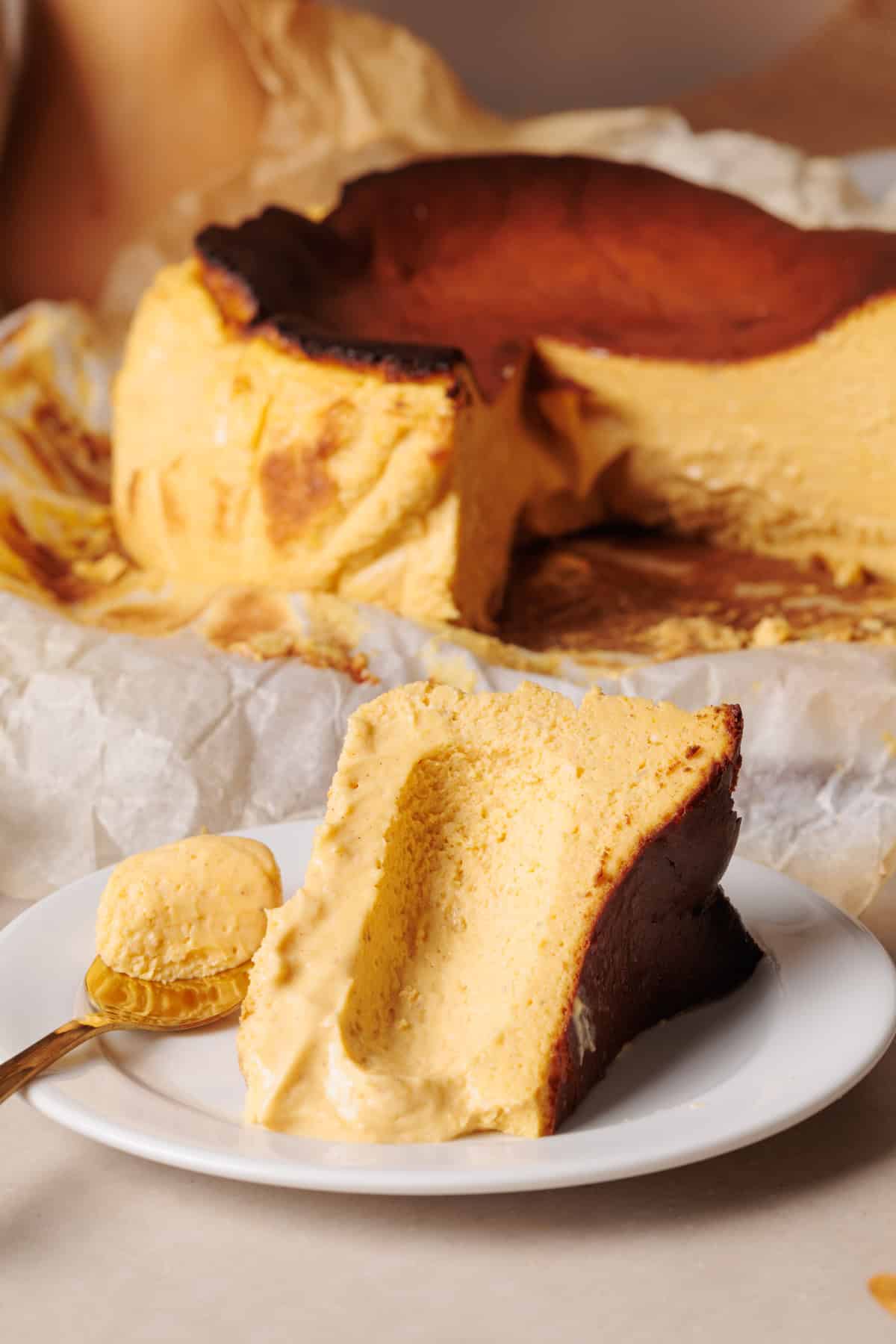 A slice of creamy, golden-brown pumpkin Basque cheesecake sits on a white plate, with a small spoonful beside it. The whole cheesecake, showing a caramelized top, is in the background on parchment paper.