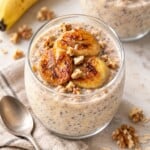 A glass jar of banana overnight oats topped with seared banana slices and walnuts, with a spoon nearby.