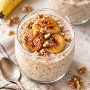 A glass jar of banana overnight oats topped with seared banana slices and walnuts, with a spoon nearby.
