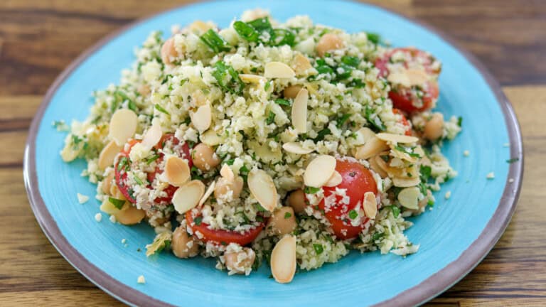 A blue plate with cauliflower tabbouleh, featuring cherry tomatoes, chickpeas, parsley, and sliced almonds on a wooden table.